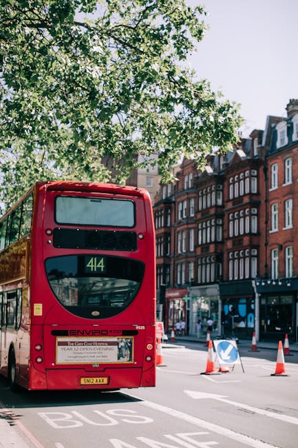 A red double-decker bus driver is stopped at a bus lane on a busy street in Maida Vale, London, with historic red-brick residential buildings featuring bay windows and decorative cornices lining the background. The bus, displaying route number 414, is positioned near the curb, with trees casting shade across the scene and traffic cones placed nearby to manage vehicle flow. The environment appears bright and clear, with sunlight illuminating the scene. This setting depicts typical urban transit and transportation elements that could be associated with house removals or logistics planning in the Maida Vale area, supporting services like furniture transport, packing, and loading during home relocation processes. Man and a Van Maida Vale, a company specializing in removals, may use such routes for efficient movement of household items between properties.