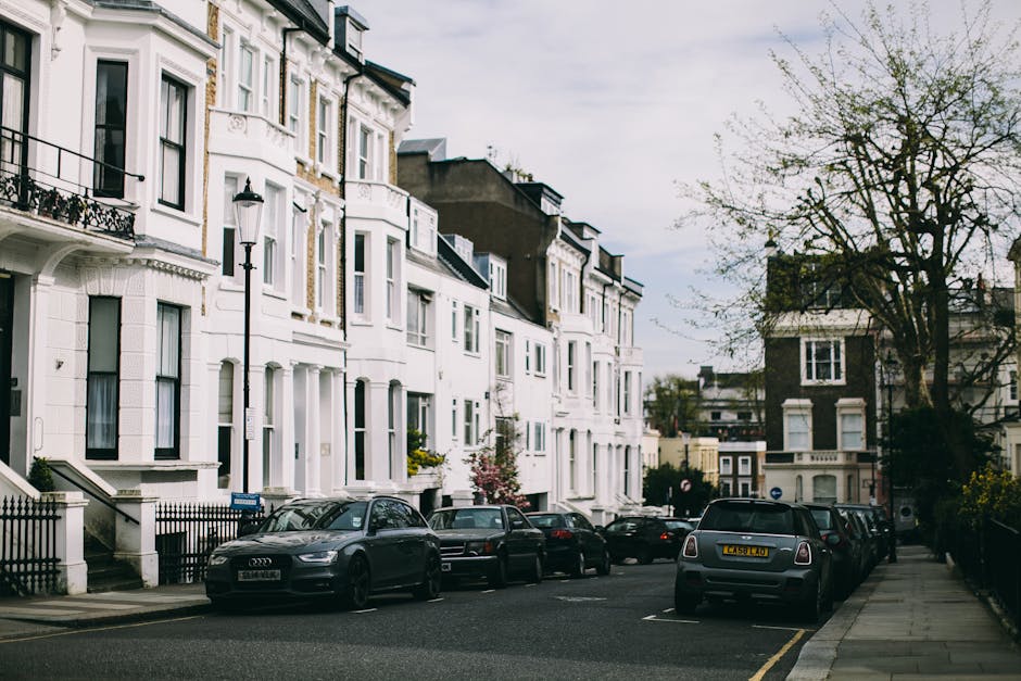 A row of white, Victorian-style terraced houses with decorative window trims and small front gardens, situated along a residential street. Several parked cars, including a black Audi and a Mini Cooper, are aligned along the curbside, with a narrow pavement on the right. In the foreground, a man and a woman, both dressed in casual clothing, are seen loading cardboard boxes and wrapped furniture pieces into a van parked in front of the house, part of a home removal process organized by Man and a Van Maida Vale. The street is lined with mature trees, some with budding leaves, and the sky is overcast, indicating a typical day for packing, moving, and furniture transport services. This scene captures the logistical aspects of house removals, with furniture blankets, packing materials, and equipment possibly visible as the team facilitates a smooth relocation within the MAIDA VALE area.
