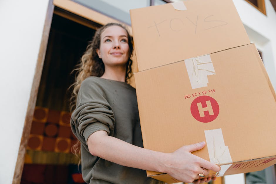 A woman with curly brown hair, dressed in a grey long-sleeve top, is holding a large cardboard box labeled 'toys' in black marker and several smaller packed boxes, inside a home during a house removal process. The boxes are secured with brown packing tape and are stacked, ready for transport. The background shows part of the indoor environment with a wooden doorway and some decorative elements on the wall. The woman is smiling slightly and appears to be preparing for a home relocation, supported by a professional removals service, such as Man and a Van Maida Vale, who is handling packing and moving logistics. The scene illustrates the packing stage of furniture transport and moving services, with attention to careful handling of packed items inside the property, just before loading into a removal vehicle.