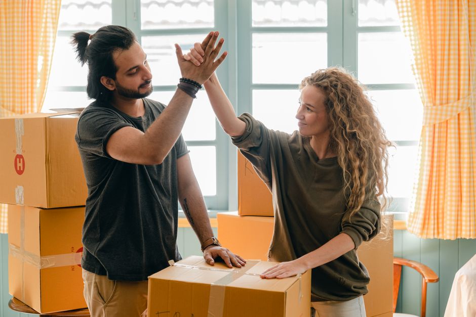 Inside a residential property, two removal workers from Man and a Van Maida Vale are engaged in a home relocation process, sharing a high-five gesture that signifies successful coordination. The room is brightly lit by natural daylight coming through a large window with light blue frames, adjacent to yellow checkered curtains that are partially drawn back. Several cardboard boxes, sealed with packing tape, are stacked on the floor and a table, indicating packing and moving preparations. One worker, with dark hair tied back and wearing a dark T-shirt, stands beside a box, while the other, with long curly hair, gestures towards him with a smile, suggesting teamwork during furniture transport or packing. The background includes a light-colored wall and the interior environment appears well-organized for a smooth loading process, reflecting professional removal services. Man and a Van Maida Vale's branding is subtly present, supporting the context of house removals and relocation services.