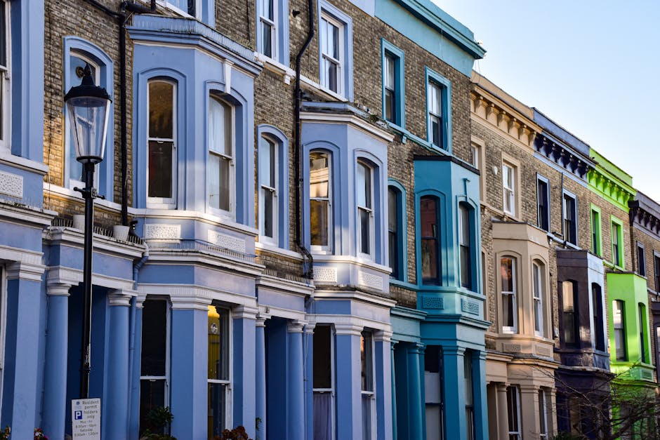 Photograph of a row of multi-storey residential buildings with Victorian-style architecture, featuring large bay windows and detailed cornices. The buildings are painted in various pastel shades including light blue, teal, beige, and light green, with some sections having brick facades. In the foreground, a black street lamp is visible on the pavement below. The image was taken during daylight with clear weather, highlighting the urban environment typical of Maida Vale. This setting reflects the premises involved in house removals and furniture transport, with the buildings serving as potential locations for packing, home relocation, and loading processes. The scene emphasizes the exterior environment often associated with professional removal services, such as those offered by Man and a Van Maida Vale, which may include loading furniture onto vans outside these properties in preparation for moves or deliveries.