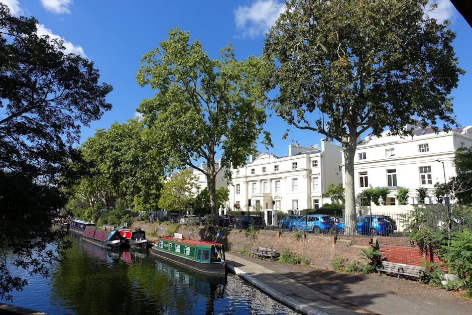 A daytime view of a residential street along a narrow waterway with several canal boats moored, including a boat with a black and white striped roof. The scene features lush green trees with full foliage, casting shadows on the pavement and boats below. Behind the trees, there are white, multi-storey terraced houses with large windows, some with small front gardens and iron railings. Parked cars, mostly in shades of blue and grey, line the street in front of the houses. The bright blue sky with a few scattered clouds suggests clear weather. In the foreground, a paved walkway runs parallel to the canal, with a low brick wall separating the path from the water. The image captures a peaceful urban environment, suitable for house removals or moving logistics, as it highlights the proximity of houses to the canal for potential boat transport services, with [COMPANY_NAME] possibly involved in local relocation or furniture transport activities.
