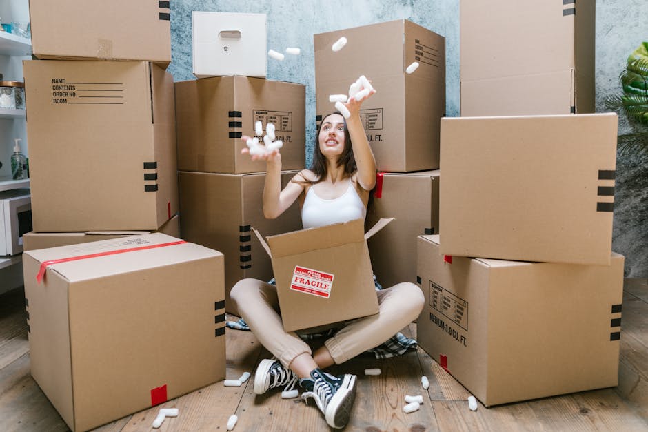 A young woman sitting cross-legged on a wooden floor inside a home, surrounded by stacked cardboard moving boxes of various sizes, some sealed with red tape and others labeled with black markings. She is wearing casual clothing and sneakers, holding a handful of packing foam pieces, which she is tossing into the air. More packing materials are scattered around her, including foam filler and shredded paper. The background shows a light-colored wall and a small potted plant in the corner, indicating an indoor space prepared for a home relocation. The scene captures the packing process involved in a furniture transport or house removal, with a focus on organized packing activities, typical of professional movers like Man and a Van Maida Vale as they prepare for a moving service near Little Venice, W9. The overall setting emphasizes the logistical stage of a packing and loading process as part of a house removal service.