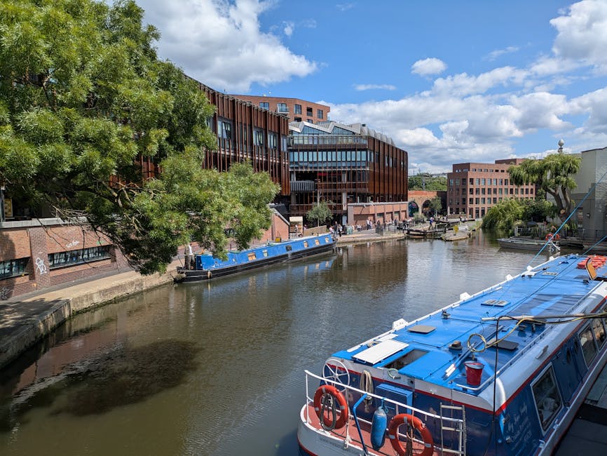 A daytime view of a canal in Maida Vale with nearby modern buildings featuring large glass windows and red brick facades, alongside lush green trees lining the water's edge. Several boats are moored along the canal, including a blue narrowboat and a larger blue and white passenger vessel in the foreground, which is docked near the pavement. The sky is partly cloudy with bright sunlight illuminating the scene. The environment suggests a lively urban area with potential activities related to house or office removals, as [COMPANY_NAME] might be involved in logistics such as furniture transport or packing during a relocation process, with the canal serving as a notable landmark in the Maida Vale district.