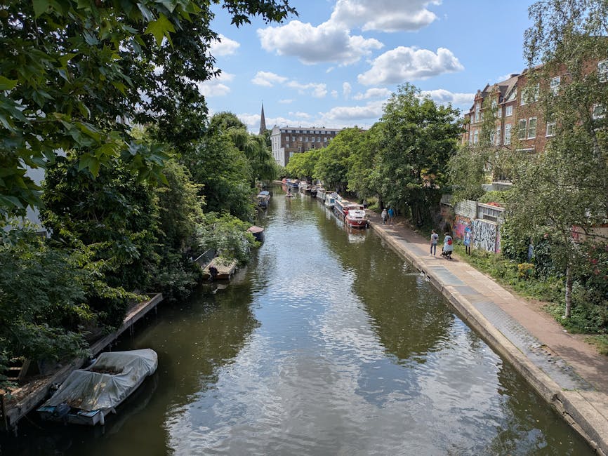 A daytime view of a canal in Maida Vale with nearby modern buildings featuring large glass windows and red brick facades, alongside lush green trees lining the water's edge. Several boats are moored along the canal, including a blue narrowboat and a larger blue and white passenger vessel in the foreground, which is docked near the pavement. The sky is partly cloudy with bright sunlight illuminating the scene. The environment suggests a lively urban area with potential activities related to house or office removals, as [COMPANY_NAME] might be involved in logistics such as furniture transport or packing during a relocation process, with the canal serving as a notable landmark in the Maida Vale district.