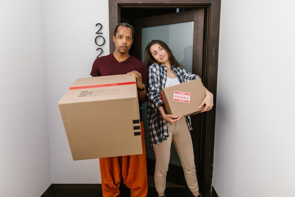 Two individuals stand inside an entryway at a residential property, each holding cardboard boxes prepared for house removals. The man on the left has a medium skin tone, is wearing a maroon T-shirt and orange trousers, and holds a large cardboard box with red and black labels. The woman on the right has fair skin, long brown hair, and is dressed in a white top with a checkered shirt over it and beige trousers; she carries a smaller cardboard box marked with 'FRAGILE' in red. They are positioned next to a dark wooden door with the house number '22' mounted vertically beside it. The interior environment features plain white walls and a black floor, with ample natural or artificial lighting illuminating the scene. The context suggests they are engaged in packing or loading activities related to moving processes, supported by the presence of packing materials and boxes, typical of home relocation services offered by Man and a Van Maida Vale, as seen in the background.