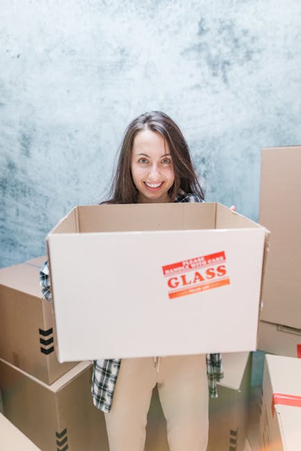 A young woman with long brown hair smiling while holding a large empty cardboard box labeled 'FRAGILE GLASS' in black and red print, standing indoors against a textured light blue wall. She is dressed in casual clothing with a plaid shirt and beige pants. Surrounding her are additional unpacked cardboard boxes of various sizes, some with packaging tape and black arrow symbols, likely part of a home relocation or packing process. The scene depicts packing and moving activities associated with house removals, as the woman prepares for or during a professional moving service. The setting suggests an organized interior space used for loading or storing household items before transporting them via van or other vehicle, consistent with services offered by [COMPANY_NAME] for furniture transport and house removals.