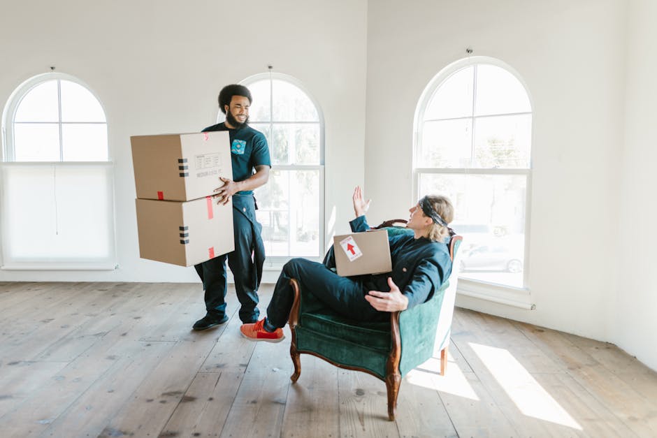 Two individuals stand inside an entryway at a residential property, each holding cardboard boxes prepared for house removals. The man on the left has a medium skin tone, is wearing a maroon T-shirt and orange trousers, and holds a large cardboard box with red and black labels. The woman on the right has fair skin, long brown hair, and is dressed in a white top with a checkered shirt over it and beige trousers; she carries a smaller cardboard box marked with 'FRAGILE' in red. They are positioned next to a dark wooden door with the house number '22' mounted vertically beside it. The interior environment features plain white walls and a black floor, with ample natural or artificial lighting illuminating the scene. The context suggests they are engaged in packing or loading activities related to moving processes, supported by the presence of packing materials and boxes, typical of home relocation services offered by Man and a Van Maida Vale, as seen in the background.