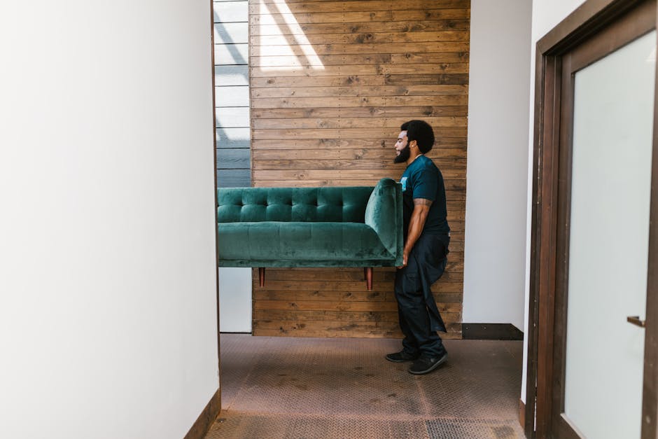 A man with a beard and dark curly hair, wearing a blue uniform, is lifting a green upholstered sofa through an interior doorway. The sofa, which features button-tufted fabric and wooden legs, is being carried into or out of a room with a wooden accent wall composed of horizontal planks. The room has a carpeted floor, and natural light is shining through a window or skylight, casting shadows on the wall. The door frame and surrounding walls are painted white, contrasting with the warm tones of the wooden wall. The scene depicts a home relocation process involving furniture transport, aligned with professional removals services provided by Man and a Van Maida Vale, as part of packing and moving logistics.