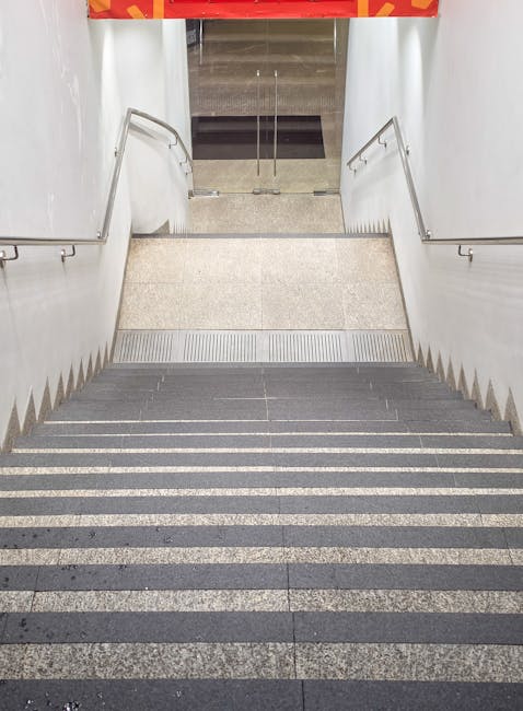 A downward view of a staircase leading to an underground or basement level, featuring dark grey non-slip treads with lighter grey anti-slip strips on each step. The staircase is flanked by white, smooth wall panels with metal handrails on both sides. At the top of the stairs, there is a small landing area with a beige, textured floor, and beyond that, a glass door with a metal frame, revealing a glimpse of an indoor environment possibly used for home relocation purposes. The space is well-lit with evenly distributed lighting, and the photo captures the interior environment prepared for moving or furniture transport, consistent with services provided by Man and a Van Maida Vale. This scene illustrates the typical stair access encountered during house removals or furniture transport, emphasizing careful navigation and staircase setup for efficient loading or unloading.