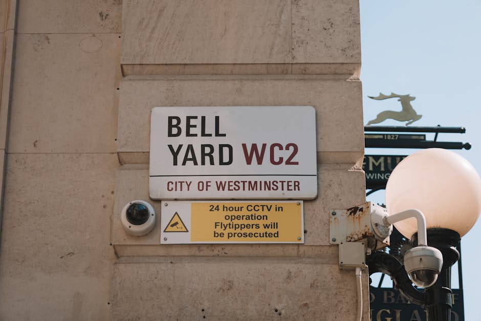 Close-up view of a street sign attached to the exterior wall of a building in Maida Vale, Westminster, indicating 'BELL YARD WC2' in black and red text, with additional signage below stating '24 hour CCTV in operation, Flytippers will be prosecuted' in yellow and black. The sign is mounted beside a security camera and a street lamp with a round, white globe shade. Part of a decorative street sign with a reindeer figure and black metal brackets is visible in the background. The building's facade is made of beige stone blocks, and the photograph is taken during daytime with natural light illuminating the scene, reflecting the urban environment where house removals and furniture transport are carried out according to local parking regulations.