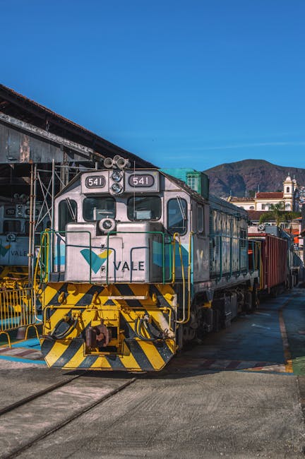 A vintage diesel locomotive painted in light blue with yellow and black striped safety markings at the front, situated on railway tracks outdoors. The engine displays the number 641 on the front and has the logo of Vale. Behind the locomotive, there are red freight containers visible, indicating a freight or cargo train. The scene is set against a backdrop of mountains and a clear blue sky. The location appears to be a rail yard or station, with some basic infrastructure and a distant building with a tower visible. This setting is relevant to house removals and moving logistics, as it illustrates the transportation process involving freight and cargo services, possibly coordinated by Man and a Van Maida Vale during a home relocation or furniture transport task.