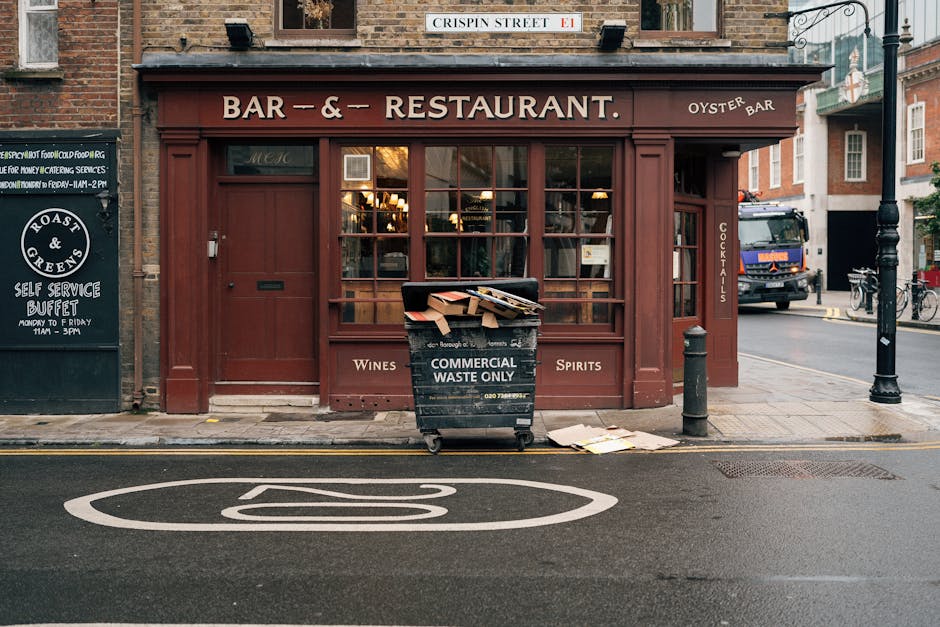 The image shows the exterior of a closed restaurant or bar on Crispen Street with a brick facade and reddish-brown wooden framing. Large windows reveal warm interior lighting, and a sign indicates it is an oyster bar serving wines and spirits. In front of the establishment, a black waste bin labeled 'Commercial Waste Only' is filled with cardboard boxes, paper, and packaging materials, some partially spilling onto the pavement. To the right, a black bollard and a lamp post are visible along the sidewalk, with a blue truck and bicycles parked further down the street. The scene captures a typical urban setting, possibly during daytime or early evening, and depicts elements related to a home relocation process, such as loading or packing, associated with professional removals services like those provided by Man and a Van Maida Vale.