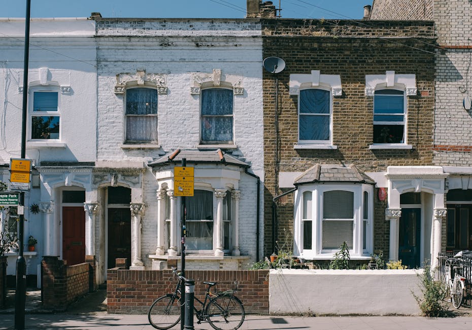 A row of terraced houses in Maida Vale with distinct architectural features, including bay windows and decorative brickwork. The leftmost house has white brickwork with two upper windows framed by stone details, while the rightmost has darker, brown brick with white window frames. In front of the houses, there are two yellow parking restriction signs mounted on a black pole, with a bicycle locked to the pole and another bicycle parked on the sidewalk. A low brick wall separates the pavement from the front garden of each property, with some potted plants visible. The scene is captured during daylight with clear skies, highlighting the urban residential environment typical for house removals and moving services, such as those offered by Man and a Van Maida Vale.